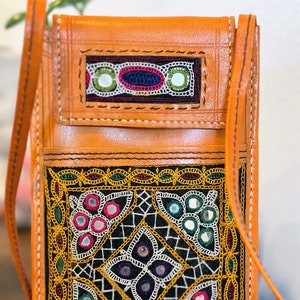 May include: Orange leather crossbody bag with intricate embroidered details. The bag features a flap closure and a long strap. The embroidery includes colorful threads and small mirrors, creating a unique and decorative design. The bag is a stylish accessory.