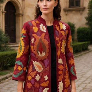 Pode incluir: Casaco bordado bordô com gola alta e mangas três-quartos. O casaco apresenta bordados florais coloridos em tons de laranja, amarelo, verde e branco. O casaco é aberto na frente e usado sobre um top preto.