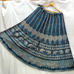 May include: A floor-length, pleated skirt in a deep teal blue color featuring a repeating pattern of white and red elephants and intricate floral motifs. The skirt is displayed on a wooden hanger and has a wide, flared silhouette with a banded waist.