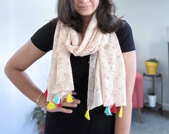 May include: A light peach-colored scarf with a floral pattern and colorful tassels. The scarf is draped around the neck. The tassels are yellow, red, and light blue.