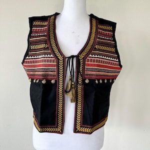 Puede incluir: Un chaleco negro sin mangas con bordados dorados y rojos. El chaleco tiene un cierre de corbata en el cuello y una borla.