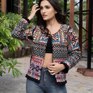 May include: A black long-sleeve jacket featuring intricate multi-colored embroidery in geometric patterns, small circular mirrors, and a scalloped hemline with black fringe trim. The jacket is worn over a black crop top and blue denim jeans.