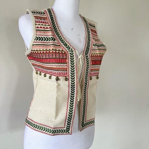 May include: A cream-colored sleeveless vest with a green and red embroidered pattern and gold-colored embellishments. The vest has a tie closure at the neck.