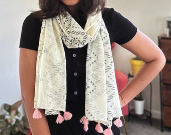 May include: A white scarf with a geometric pattern and pink tassels. The scarf is worn around the neck of a person wearing a black shirt.