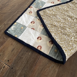 May include: A patchwork quilt with a blue, white, and gold pattern featuring angels and stars. The quilt is folded in half, revealing a floral patterned fabric on the back.