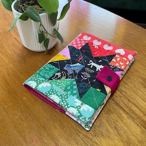 May include: A patchwork notebook cover with a star design. The cover is made from various patterned fabrics in pink, green, black, and red. A magenta strap with a black button fastens the cover. A potted plant is in the background.