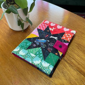 May include: A patchwork book cover with a starburst design. It features a variety of fabric patterns, including hearts, geometric shapes, and roller skates. The cover has a pink button closure and a contrasting pink interior.