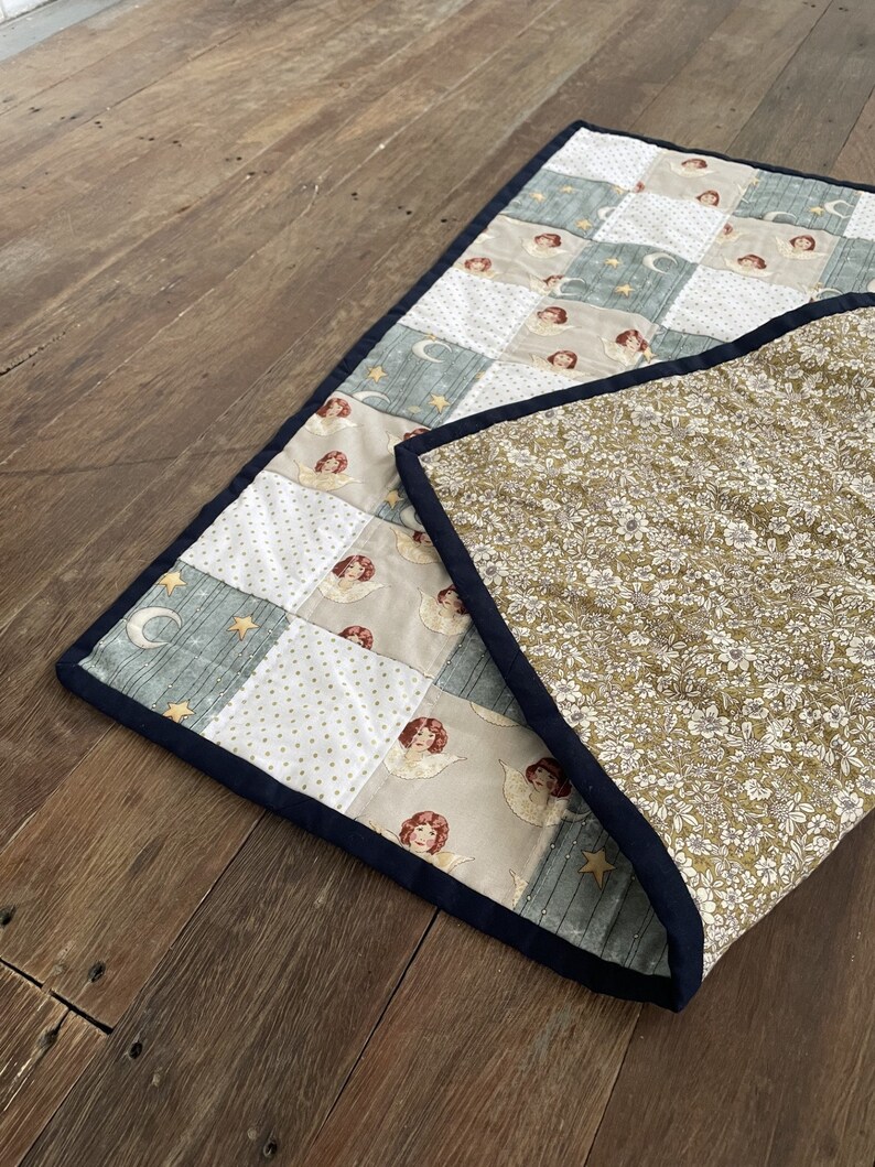 May include: A patchwork quilt with a blue, white, and gold pattern featuring angels and stars. The quilt is folded in half, revealing a floral patterned fabric on the back.