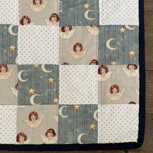 May include: A patchwork quilt with a blue, white, and beige color scheme. The quilt features a repeating pattern of angels and stars. The quilt is edged with a dark blue trim.