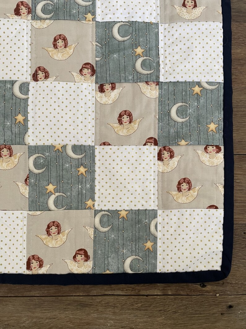 May include: A patchwork quilt with a blue, white, and beige color scheme. The quilt features a repeating pattern of angels and stars. The quilt is edged with a dark blue trim.