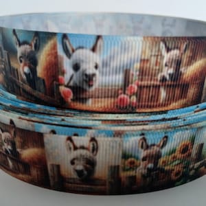 May include: A roll of ribbon featuring a repeating pattern of donkeys in a farm setting. The ribbon has a blue sky background and is decorated with sunflowers, fences, and red flowers. The ribbon is likely used for crafts or gift wrapping.