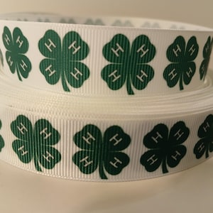 May include: White ribbon with a repeating pattern of green four-leaf clovers, each with the letter "H" inside. The ribbon is rolled into two stacks, showing the pattern clearly.