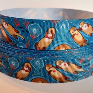May include: A roll of teal ribbon featuring a repeating pattern of cartoon otters in various poses. The otters are brown with red hearts and are set against a background of white and red sea life.