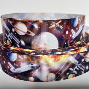 Könnte beinhalten: Gerolltes Band mit Weltraum-Motiv. Das Band zeigt Planeten, Raumschiffe und Sterne auf dunklem Hintergrund mit orangefarbenen und gelben Akzenten. Geeignet für Bastelarbeiten oder Geschenkverpackungen.