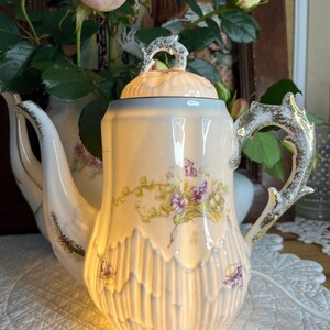 Könnte beinhalten: Cremefarbene Porzellan-Teekanne mit einem Blumendesign mit lila Blumen und grünen Blättern. Die Teekanne hat einen dekorativen Griff und Ausguss mit goldenen Akzenten. Der Deckel ist mit einem dekorativen Griff versehen. Die Teekanne wird von innen beleuchtet und erzeugt ein warmes Leuchten.