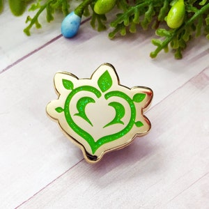 Op de afbeelding: Gouden emaille pin met een groen glitterhart ontwerp. Het hart is omgeven door groene bladeren.