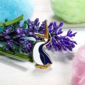 May include: A gold-tone enamel pin of a cartoon bird with a white body, blue wings, and a long orange beak. The bird is looking up and has its mouth open.