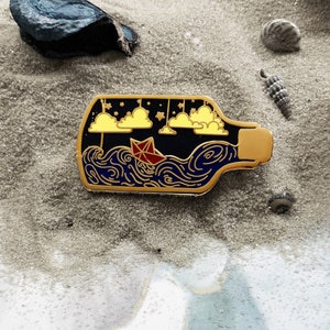 Pode incluir: Pin de esmalte dourado que representa uma garrafa de vidro com uma rolha de cortiça. A garrafa contém um oceano azul com nuvens brancas e um barco de papel vermelho.
