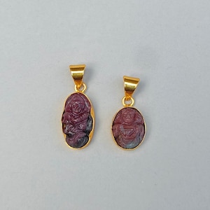 Könnte beinhalten: Zwei goldbeschichtete Anhänger mit geschnitzten rubinroten Figuren. Die Anhänger haben eine goldene Öse zum Aufhängen. Die Figuren sind mit komplizierten Details geschnitzt. Die Anhänger sind oval und leicht unterschiedlich groß.