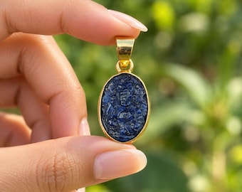 May include: An oval-shaped pendant featuring a deep blue carved gemstone set in a polished gold-toned metal frame. The stone displays intricate relief carvings of a deity figure. A small gold-toned bail is attached to the top of the pendant.