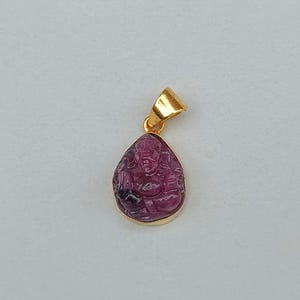 May include: A gold pendant with a carved, teardrop-shaped, pink gemstone. The gemstone features a detailed carving of a figure. The pendant has a gold bail for attaching to a chain. The background is a solid, light gray.