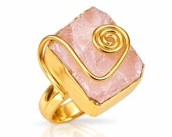 May include: A gold-toned ring featuring a large, square-cut, pale pink raw crystal stone. The stone is secured within a gold-colored metal setting and accented with a decorative gold wire spiral design that sits atop the surface of the crystal.
