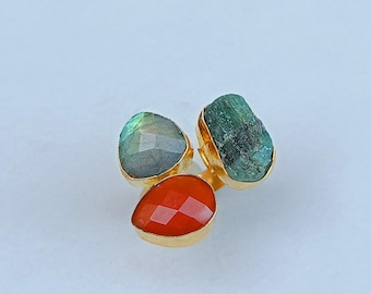 May include: A gold ring featuring three distinct gemstones: a faceted grey-blue labradorite, a rough-textured green emerald, and a smooth, faceted orange carnelian, all set in a gold-toned metal band against a plain light background.