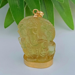 Może przedstawiać: Żółty wisiorek z kamienia szlachetnego wyrzeźbiony w kształcie słonia. Wisiorek ma złoty uchwyt i złotą podstawę. Rzeźba słonia jest szczegółowa, z widocznymi kłami, trąbą i uszami. Tło jest rozmyte z zielonymi liśćmi.