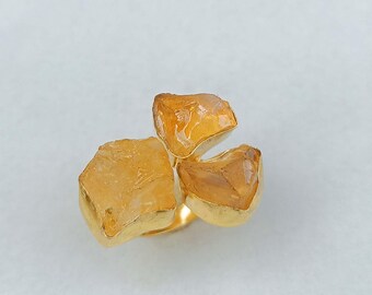 May include: A gold-toned ring featuring three rough-cut, amber-colored gemstones. The gemstones are set in a gold band, creating a unique and naturalistic design. The ring's design is reminiscent of a floral arrangement.
