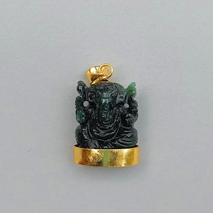 Puede incluir: Colgante tallado en verde oscuro que representa a la deidad hindú Ganesha. El colgante está engastado en una base y un asa de metal dorado. La figura está sentada con detalles intrincados, mostrando la cabeza de elefante y múltiples brazos. Para collar.