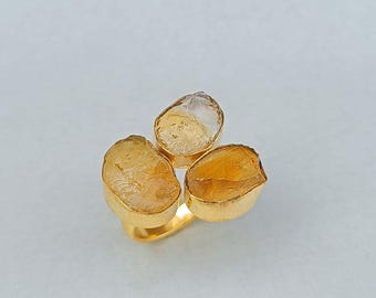 May include: A gold ring featuring three raw, golden-yellow citrine stones. The stones are set in individual gold bezels, arranged in a cluster. The ring's band is also gold, creating a cohesive and elegant design. The citrine stones have a natural, organic shape.