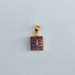 Pode incluir: Um pingente de ouro com uma pedra roxa quadrada com um rosto esculpido.