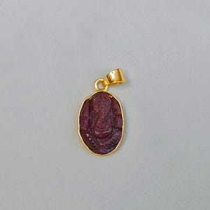 May include: A gold-colored pendant with a carved, maroon-colored figure. The pendant has a gold bail for a chain. The figure is an oval shape with a detailed carving of a deity.