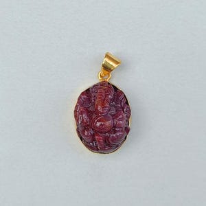 Puede incluir: Un colgante de color rubí con una imagen tallada de la deidad hindú Ganesha. El colgante tiene una fianza dorada y un marco dorado alrededor del rubí de forma ovalada. La talla es detallada e intrincada.