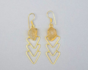 May include: Gold earrings with a geometric design featuring three stacked triangles. Each earring is topped with a light brown, irregularly shaped gemstone.