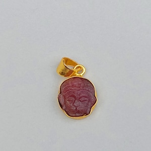 Op de afbeelding: Een goudkleurige hanger met een gesneden, rode edelsteen die een gezicht afbeeldt. De hanger heeft een lus aan de bovenkant voor een ketting. De ovaalvormige edelsteen is gezet in een gouden bezel.