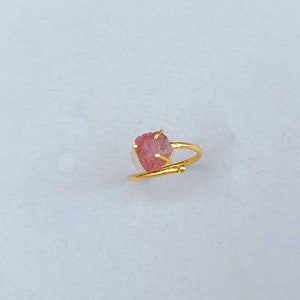 May include: A gold-colored ring featuring a raw, pink gemstone. The gemstone is irregularly shaped and set with small prongs. The ring band curves around the finger, with a small decorative element near the gemstone.