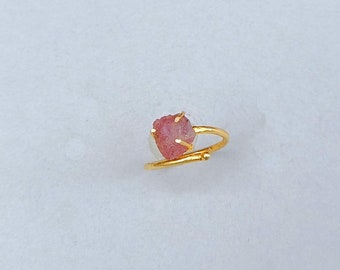 May include: A gold-colored ring featuring a raw, pink gemstone. The gemstone is irregularly shaped and set with small prongs. The ring band curves around the finger, with a small decorative element near the gemstone.