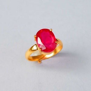 May include: A gold ring with a large, oval-shaped ruby gemstone. The ruby is a deep red color and is set in a simple, four-prong setting.