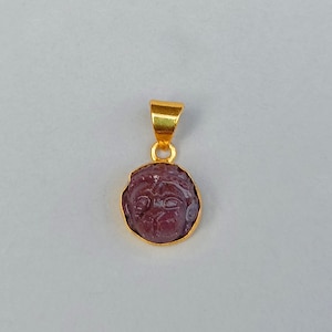 May include: A gold pendant featuring a carved, reddish-purple face. The face is set within a gold bezel and has a gold loop for a chain. The pendant is displayed against a light gray background.
