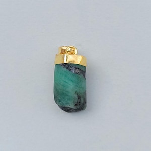 May include: A raw emerald pendant with a gold-colored cap and bail. The emerald is a vibrant green color with dark inclusions. The pendant is set against a light gray background.