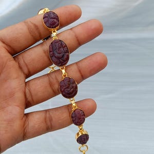 May include: A gold-tone link bracelet featuring five oval-shaped, deep red carved gemstone charms. Each charm displays a relief carving of a deity figure. The bracelet is constructed with thin gold-colored metal connectors and a toggle clasp closure.