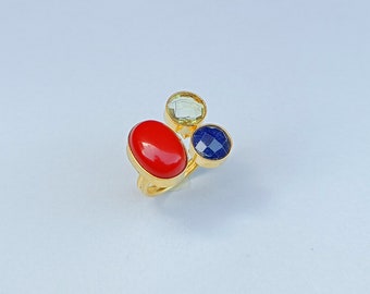 May include: A gold ring featuring three distinct stones: a large, smooth, oval-shaped red stone, a smaller, round, faceted light green stone, and a round, faceted dark blue stone, all set in a polished gold-tone metal band.