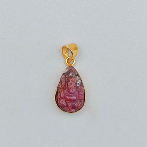 May include: A teardrop-shaped pendant with a carved red gemstone depicting a deity, set in a gold-colored frame. The pendant has a gold loop for a chain. The gemstone has a detailed carving of a figure.