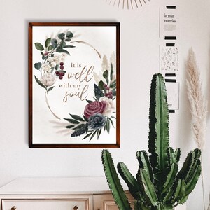 May include: A framed print featuring a watercolour floral design and the text "It is well with my soul". The print is hanging on a white wall above a white dresser with a cactus plant in a woven basket.