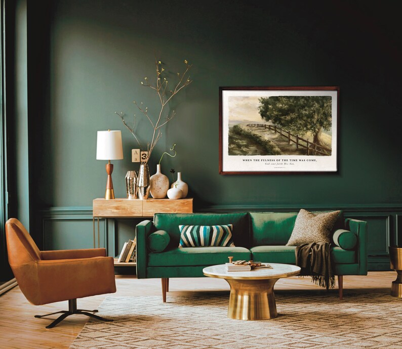 May include: A green velvet sofa with a brown throw blanket sits in a living room with a wooden console table and a gold lamp. A framed watercolor painting of a path through a forest hangs on the wall above the sofa. The painting includes the text "When the fullness of the time was come."