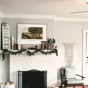 May include: A white brick fireplace with a black metal fireplace screen. Above the fireplace is a framed print of a landscape with a path leading to a wooden fence. The print is surrounded by greenery and a wooden sign with the text "In all things give thanks".