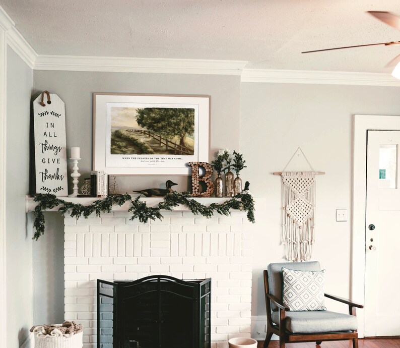 May include: A white brick fireplace with a black metal fireplace screen. Above the fireplace is a framed print of a landscape with a path leading to a wooden fence. The print is surrounded by greenery and a wooden sign with the text "In all things give thanks".