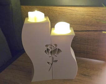 Könnte beinhalten: Ein weißer Kerzenhalter mit Rosenmuster, der zwei brennende Kerzen hält. Der Halter hat eine geschwungene Form und besteht aus einem festen Material. Die Kerzen strahlen ein warmes, gelbes Licht aus und erzeugen eine gemütliche Atmosphäre.