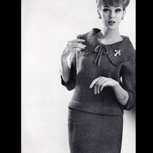 May include: A black and white photo of a woman wearing a knitted dress with a large collar and a bow tie. The dress has a textured pattern and the woman is wearing a brooch on the collar.